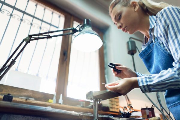Kvinna i blåa kläder håller i ett föremål och tittar på det med förstoringsglas för att kunna värdera det.