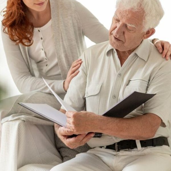 En yngre kvinne sitter bredvid och håller armen om en äldre man som håller i en pärm med papper. Kvinnan hjälper mannen med bodelning efter ett dödsfall