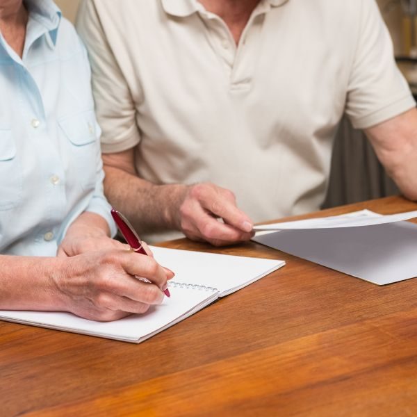 Två personer sitter ner vid ett bord och antecknar med penna och papper för att göra en dödsboanmälan.